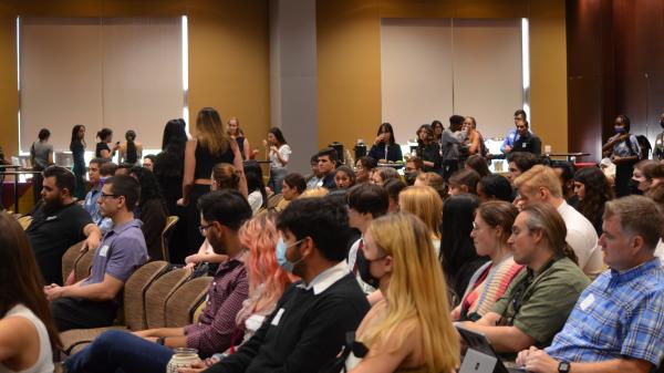 A crowded conference room with attendees seated and some standing, engaging in conversations. The atmosphere appears formal, with some participants using laptops and wearing name tags.