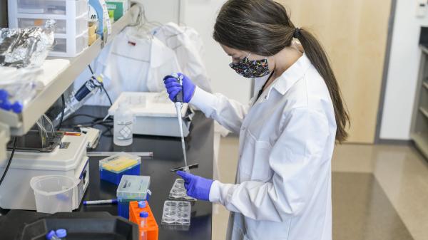 A woman in a white lab coat and mask works with a pipette in a laboratory. The workspace contains various lab equipment, including plastic containers and sample trays.