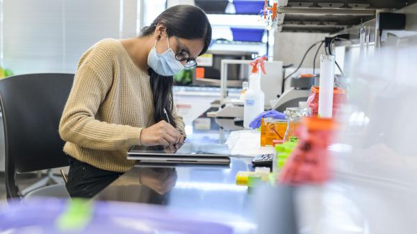 A woman wearing a beige sweater, glasses, and a face mask is writing on a tablet or digital device in a laboratory setting. The background shows blinds and a window, suggesting the lab is situated in a well-lit area.