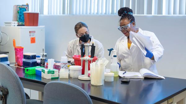 Two women are working in a laboratory setting. Both are wearing lab coats, safety glasses, and face masks.