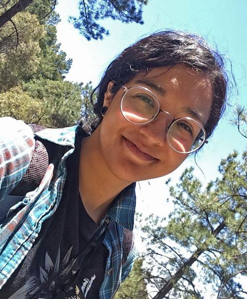 A woman in glasses smiling in front of pine trees.