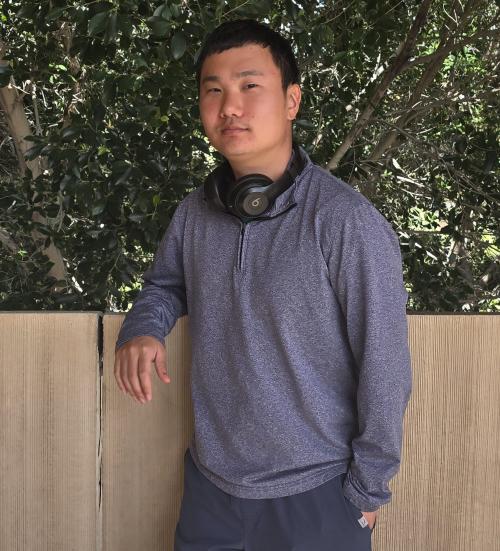 A man with dark hair and headphones around his neck leans against a wall.