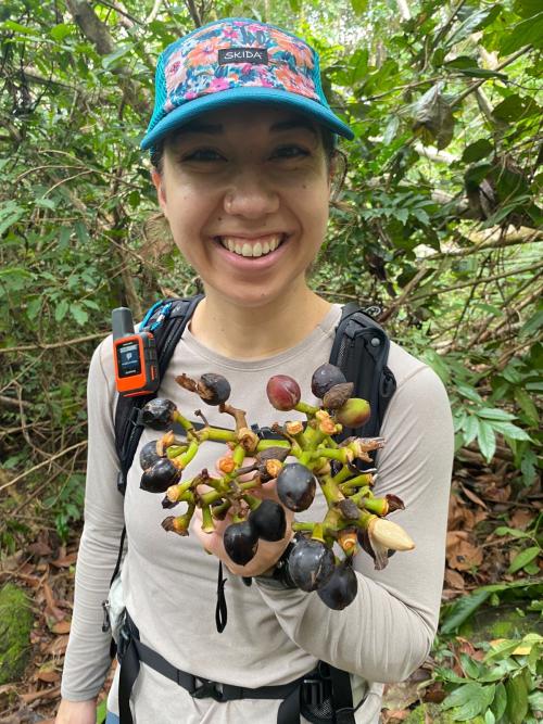 A woman in a blue hat smiling and holding up a plant in the rainforest.