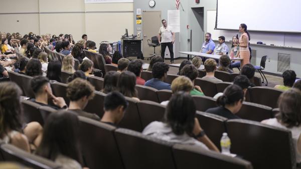 A lecture hall filled with attentive attendees. At the front, a presenter stands near a podium, while three individuals sit beside her. The setting suggests an academic or professional presentation or discussion.