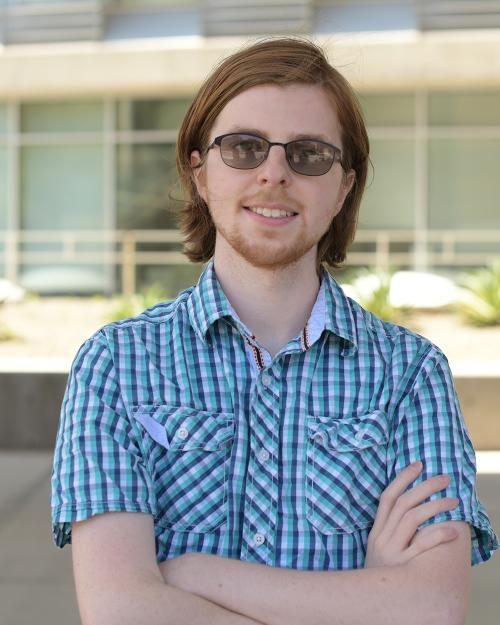 A blonde man with a goatee and sunglasses stands crossing his arms and smiling.